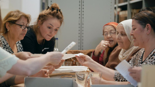 Filmstill: Mehrere Frauen sitzen an einem Tisch und betrachten gemeinsam Dokumente.