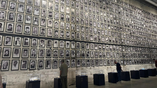 Foto: Eine hohe Wand ist dicht mit gerahmten Schwarz-Weiß-Porträts bedeckt. Zwei Personen betrachten die Bilder und Vitrinen davor.