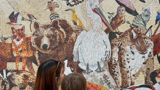 Foto: Zwei Personen stehen vor einer großen Mosaikwand, die diverse Tiere und Vögel zeigt