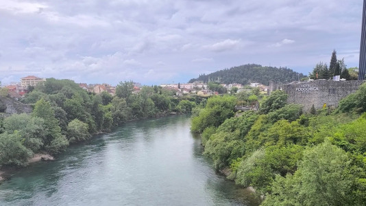 Breiter Fluss zwischen dicht bewachsenen Ufern, dahinter Stadtbebauung. Rechts Gebäude mit Schriftzug „Hotel Podgorica“, bewölkter Himmel.